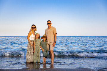A happy young family posing together at the beach by the shoreline, smiling and enjoying a sunny day by the sea, warm light, clear sky, soft sand, and gentle waves in the background