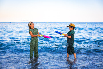 A girl and a boy playing on the seashore with colorful water pistols, splashing water, laughing and...
