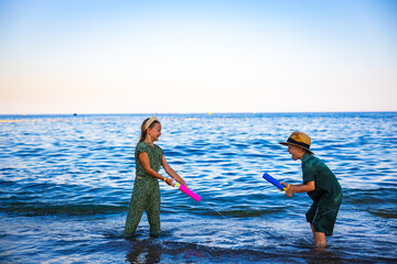 A girl and a boy playing on the seashore with colorful water pistols, splashing water, laughing and...