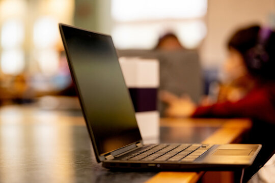 Open laptop computer on wooden table in school library study area with blurred background of students studying