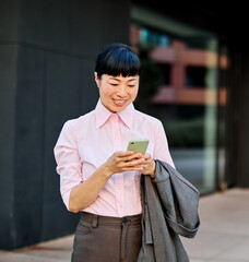 Portrait of a young asian japanese businesswoman woman using a smartphone mobile phone walking down the street, surrounded by moder corporate office buildings