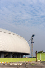 A large construction crane stands next to the curved, corrugated metal roof of a modern stadium under a partly cloudy sky. Industrial development.