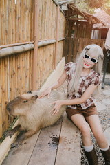Young woman in sunglasses playing with a capybara in a cozy outdoor setting with wooden fence and...