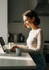 Smiling young woman working on a laptop in her kitchen, illuminated by morning light, Focused and smiling young woman working on her computer in a bright kitchen setting