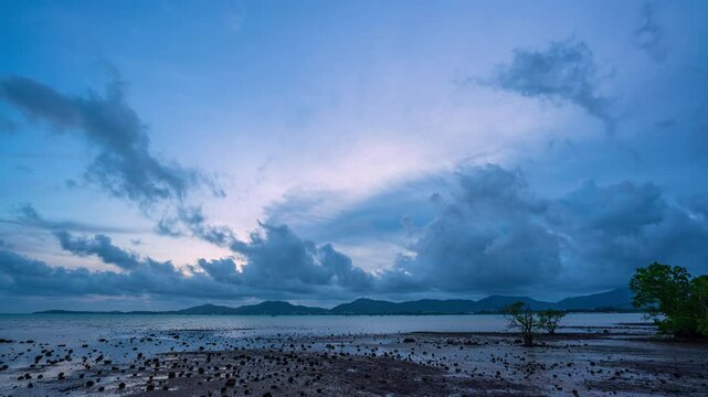Time lapse A tranquil coastal landscape showcasing a low-tide mudflat under a dramatic dusk sky, with scattered mangrove trees and distant mountains silhouetted against the setting sun. moving clouds