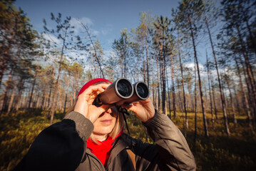 A middle-aged girl with binoculars in a red hat and a green jacket stands in the forest on a sunny...