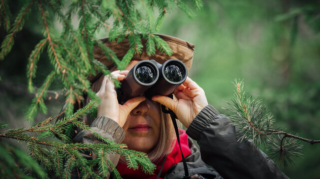 A middle-aged girl in green clothes and a green hat with a brim looks through binoculars from behind the branches of trees.