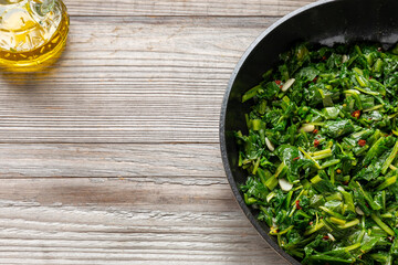 A pan with Roman-style chicory greens sautéed in olive oil (Cicoria Ripassata alla Romana). A bottle of olive oil on the side. Rustic wooden background with copy space.