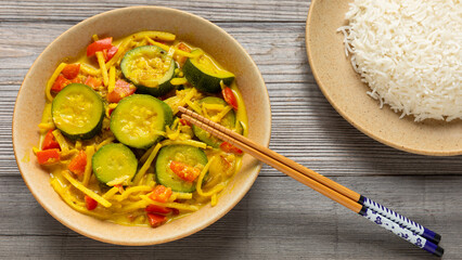 Vegan Thai yellow curry made of zucchini, red bell pepper, and bamboo shoots in a bowl. Chopsticks, rice on a plate. Rustic wooden table.