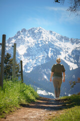 A female hiker walks along a narrow trail in spring, with the majestic Tegelberg of the Allgäu Alps rising snow-capped in the background—perfect for themes of outdoor adventure, hiking, and recreation