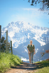 A female hiker walks along a narrow trail in spring, with the majestic Tegelberg of the Allgäu Alps rising snow-capped in the background—perfect for themes of outdoor adventure, hiking, and recreation