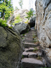 Stone steps in sandstone formation of stone city in Ostas in Czech Republic, leading up. Grey and beige rock wall, uneven steps, green trees, high contrast of light and shadow.