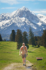 A male hiker with a vintage backpack walks along a rural field path, framed by the stunning, snow-capped peaks of the Allgäu Alps—ideal for adventure, travel, and outdoor recreation concepts.
