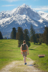 A male hiker with a vintage backpack walks along a rural field path, framed by the stunning, snow-capped peaks of the Allgäu Alps—ideal for adventure, travel, and outdoor recreation concepts.