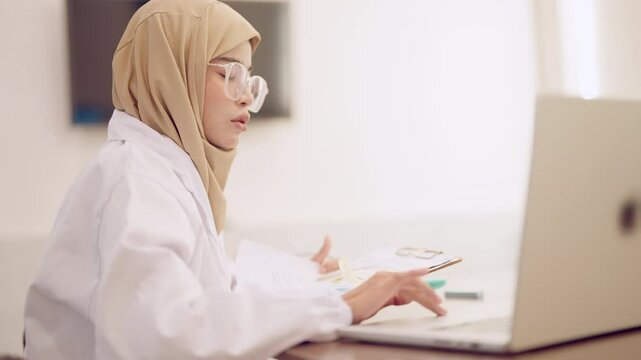 A woman wearing a lab coat is sitting at a desk with a laptop and a clipboard. She is looking at the laptop screen