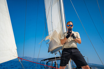 Man standing with smartphone on the bow of a sailing yacht under full sails near Croatia. Blogging,...