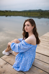 A girl in a blue dress sits on a pier on wooden planks near a lake. She hugs her legs and looks away