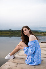 A girl in a blue dress sits on a pier on wooden planks near a lake. She hugs her legs and looks away