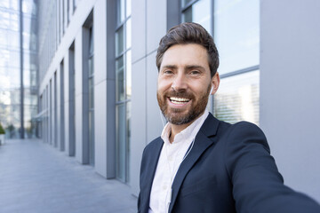 A smiling businessman in a suit takes a selfie outside a modern building, wearing earbuds.
