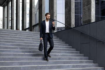 A businessman in a suit descends outdoor stairs while holding a phone and briefcase, modern city...