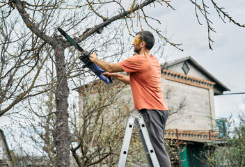 Young man cutting trees using electrical chainsaw in motion at his backyard