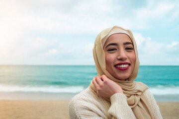 Confident young woman wearing a beige hijab and cozy sweater, smiling while standing by the sea...