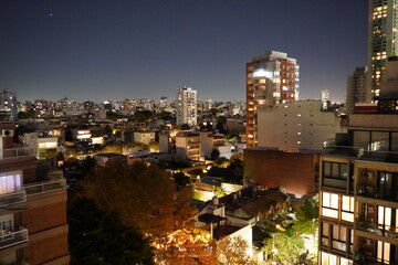 City skyline at dusk with illuminated buildings and a vibrant sunset gradient in the sky. Urban landscape featuring silhouettes of high-rise apartments and glowing city lights.