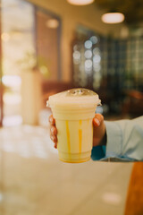 Refreshing yellow drink in clear cup held by hand in modern cafe setting with soft lighting and...