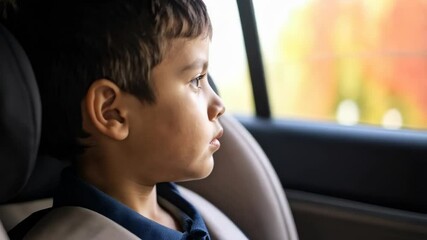 Pensive child sitting in a car seat buckled up safely looking out the window during a car ride, wearing a blue collared shirt