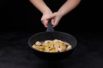 Hands holding a frying pan with seasoned chicken pieces, showcasing the cooking process against a dark background, emphasizing culinary skills and food preparation techniques