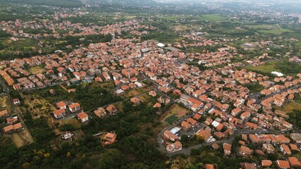 Drone photo of Zafferana Etnea, a town on the slopes of Mount Etna in Sicily, showing red-roof houses, green vegetation and a scenic volcanic landscape.