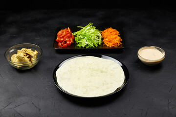 Fresh ingredients arranged for a delicious wrap, featuring a tortilla, sliced vegetables, and sauces on a dark textured background, highlighting culinary preparation and presentation