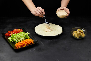 Hands of a person preparing a fresh wrap with sauce, surrounded by vibrant vegetables including lettuce, carrots, and tomatoes on a dark textured surface, showcasing culinary creativity