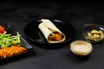 Colorful vegetable wrap on a black plate, surrounded by fresh ingredients like lettuce, carrots,...