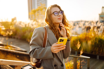 Stylish woman on roof terrace with phone in hand chatting and enjoying sunset. Young woman posing...