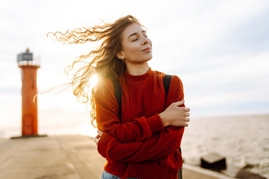 Portrait of beautiful woman on pier admiring seascape with lighthouse in background. Sweet woman feels freedom in fresh air. Freedom, nature concept, cozy atmosphere.