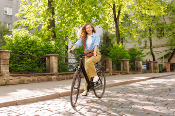 Naklejka premium Young tourist with curly hair rides a bicycle along sunny European streets. A woman in casual clothes enjoys a ride with a bicycle at sunset. Tourism concept. Active lifestyle.