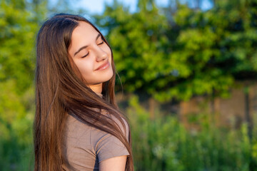 Young woman enjoying sunlight with smile. Portrait of a young woman with long hair smiling peacefully with eyes closed, basking in warm sunlight outdoors.