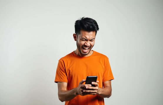 Angry Asian man in orange t-shirt looking at smartphone with message on isolated background. Frustrated male with negative emotions. Stress problem upset by bad news on social media.