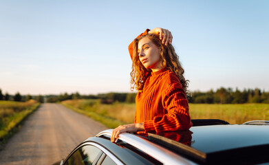 Happy woman leans out of car window and enjoys scenery from window. Young female traveler feels...