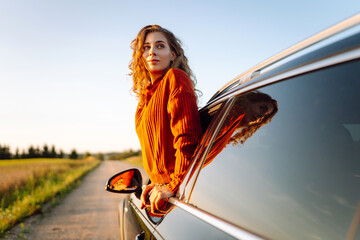 Happy woman leans out of car window and enjoys scenery from window. Young female traveler feels...