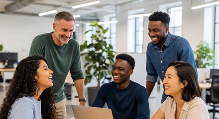 A diverse group of colleagues are gathered around a laptop in a modern office, smiling and laughing during a collaborative meeting.