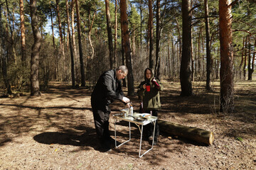 Couple engaged in early morning camping routine, with a compact cooking station set up in a sunlit pine forest, preparing food and drinks