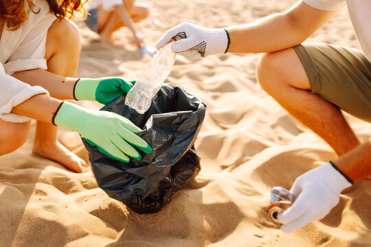 A group of volunteers collect plastic bottles and trash on a sandy beach. Young men and women collect trash on the ocean shore, clean up the environment. The concept of ecology and volunteering. - Powered by Adobe