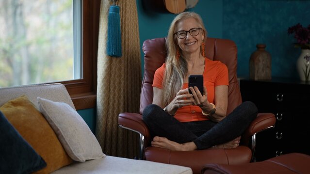 Portrait of excited surprised mature woman sits in a brown leather chair, reclining, her legs crossed, smiling broadly and looking at her smartphone. She is wearing orange shirt, black pants, glasses - Powered by Adobe