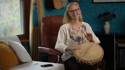 Mature woman with blonde hair and glasses smiles while playing a djembe drum. She sits in a leather chair in a room with a window, a couch, and a bookshelf.