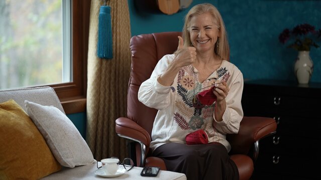 Mature woman sits in a leather chair knitting a red scarf. She smiles and gives a thumbsup, while a ball of yarn rests on her lap. A cup and eyeglasses sit on the table beside her.