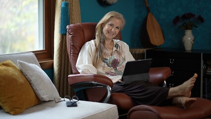 Mature woman with blonde hair sits in a leather chair with a tablet computer on her lap and her...