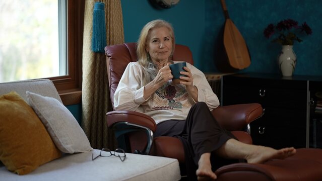 Portrait of smiling mature woman with graying hair sits in a brown leather recliner holding a blue mug and wearing ethnic blouse, black pants, and no shoes.