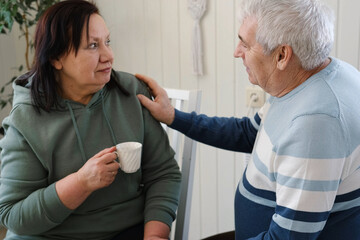 Fototapeta premium Old couple have breakfast sitting at the kitchen table, drink coffee. Embracing elderly man and woman. Senior couple together marriage in retirement talking with each other. Enjoying the pleasantries